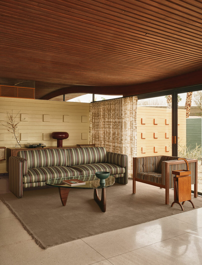 A mid-century modern living room with a striped sofa and armchair, wooden coffee table, side tables, beige rug, and floor-to-ceiling windows with patterned curtains.