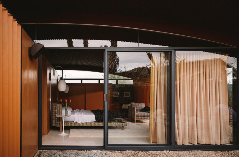 A modern room with glass sliding doors, orange walls, and beige curtains, showing a bed, armchair, tulips on a table, and outdoor gravel pathway.