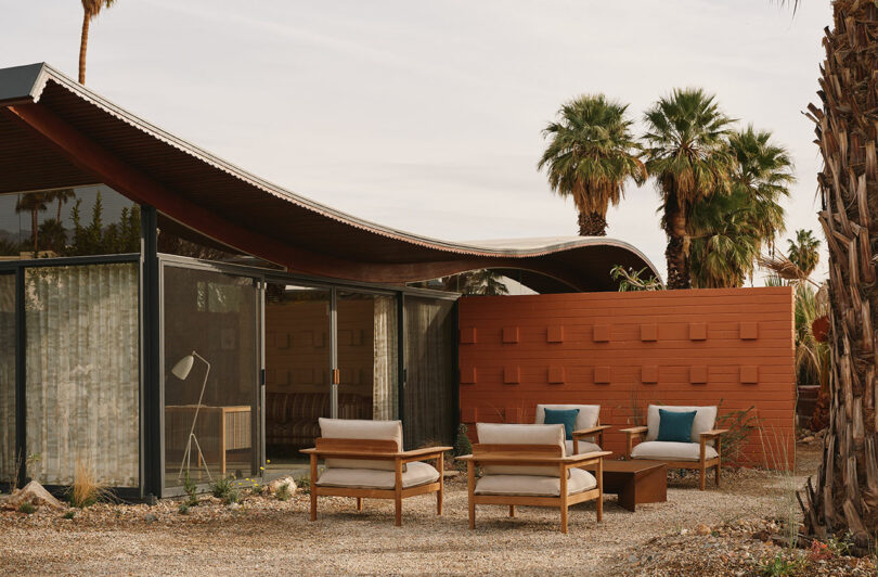 A modern house with a wavy roof, glass walls, and a patio featuring outdoor chairs and a table, surrounded by gravel, palm trees, and a red privacy wall.
