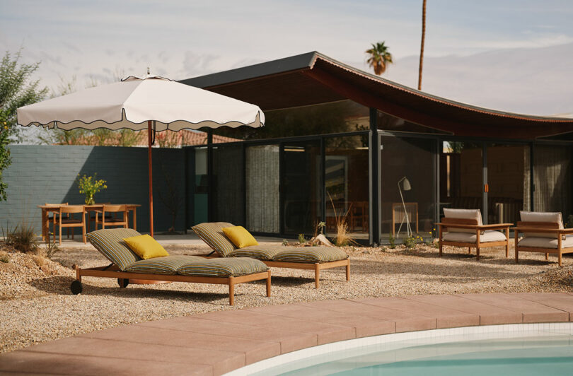 Poolside area with two lounge chairs, a white umbrella, and a modern house with large glass windows in the background. Desert landscaping surrounds the seating area.