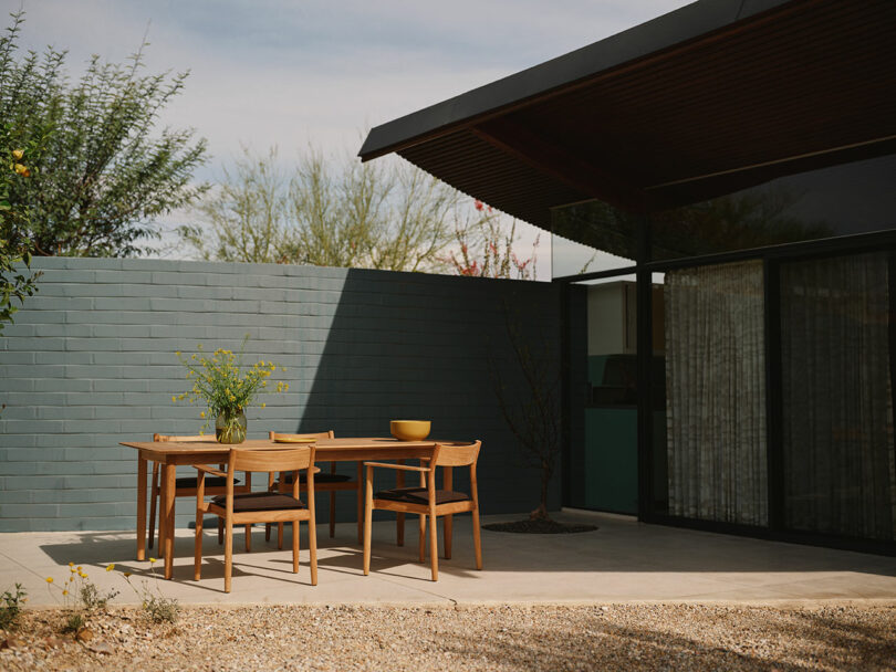 A modern outdoor patio with a wooden dining table and four chairs, set against a gray brick wall and next to a house with large glass windows.