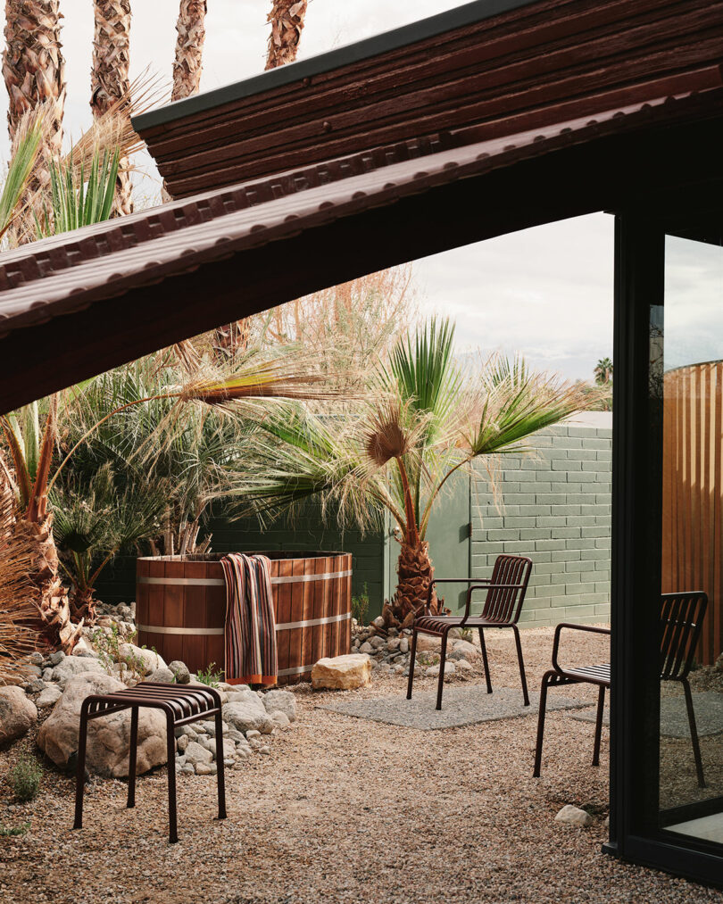 A small patio with gravel ground, metal chairs, a wooden hot tub, palm trees, and a green brick wall, partially shaded by a metal roof.