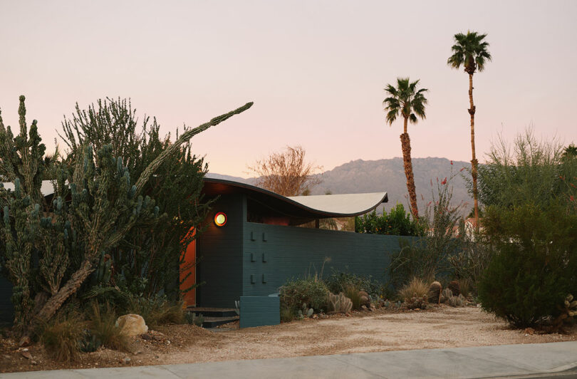 A modern, flat-roofed house with a curved overhang, surrounded by desert plants and palm trees, set against a mountainous backdrop at sunset.