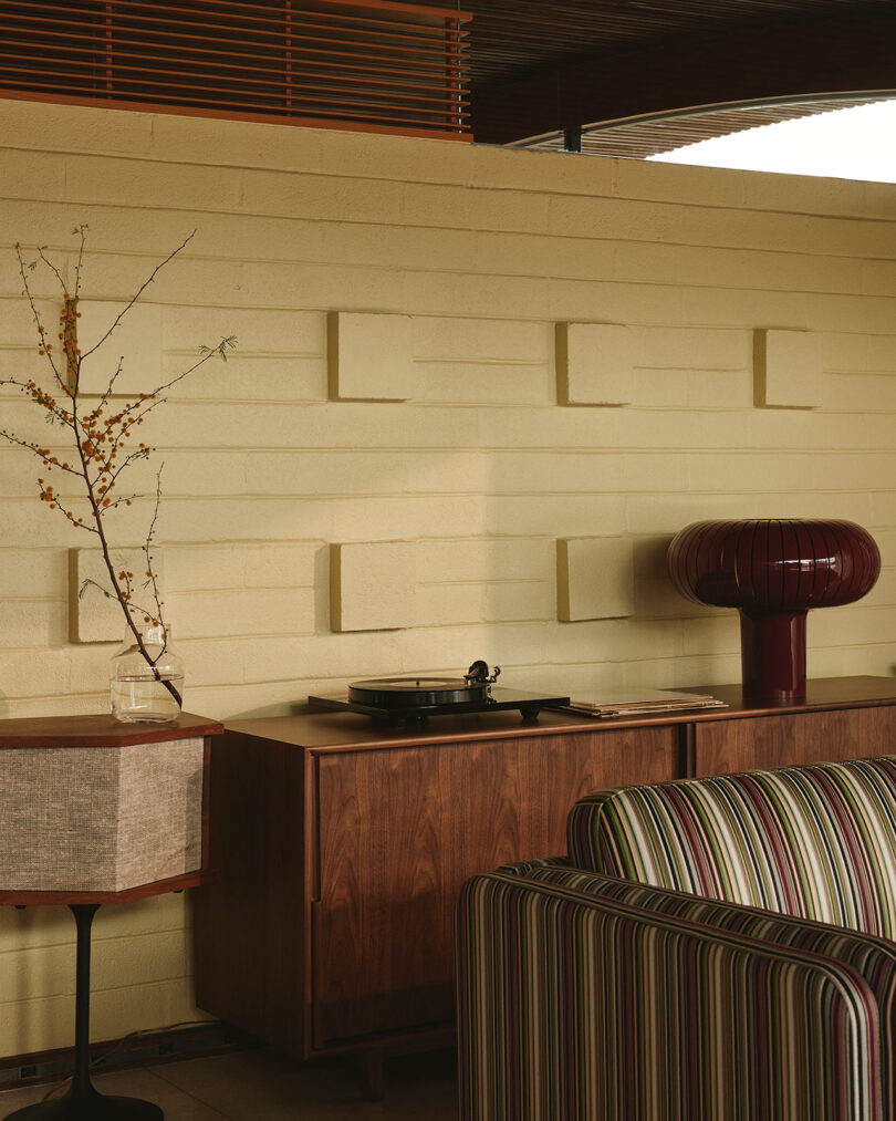 Mid-century modern interior with a striped sofa, wooden sideboard, record player, maroon lamp, vase with branches, and a textured cream wall with geometric accents.