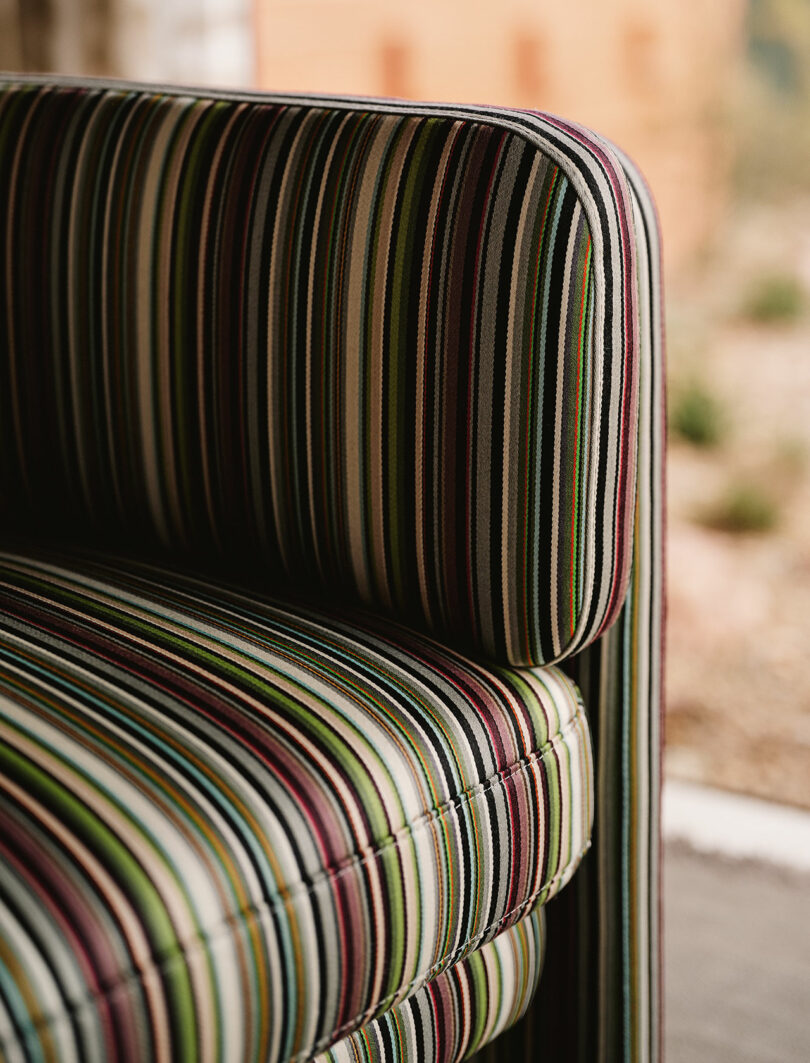Close-up of a cushioned chair with vertical multicolored stripes, featuring green, burgundy, yellow, and blue tones, in an indoor setting with blurred background.