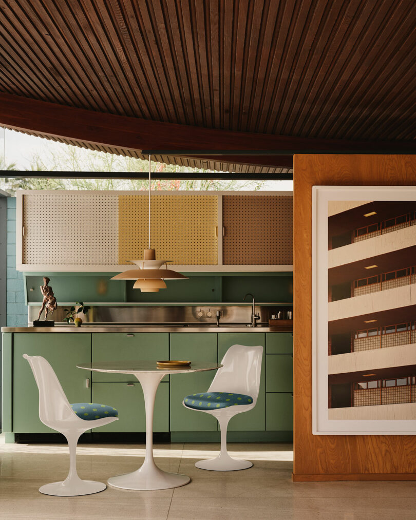 A mid-century modern kitchen with green cabinets, a white tulip table, two white chairs, a pendant light, and a large framed photo of a building on a wooden wall.