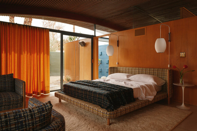 Mid-century modern bedroom with wood paneling, plaid upholstered bed, orange curtains, glass doors to an outdoor area, and two pendant lights above a white bedside table.