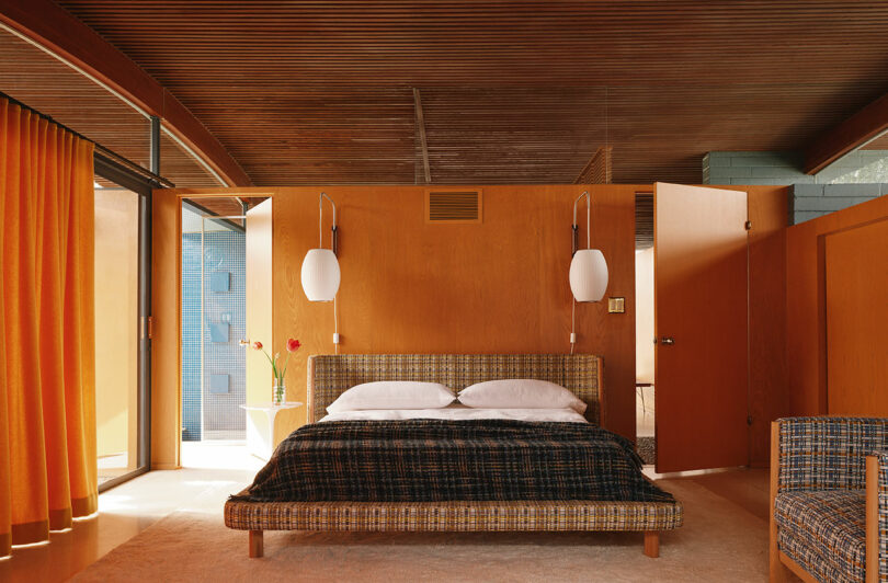 A mid-century modern bedroom with a wooden ceiling, an upholstered bed, pendant lights, orange curtains, and an open door, featuring warm, earthy tones and minimalist decor.