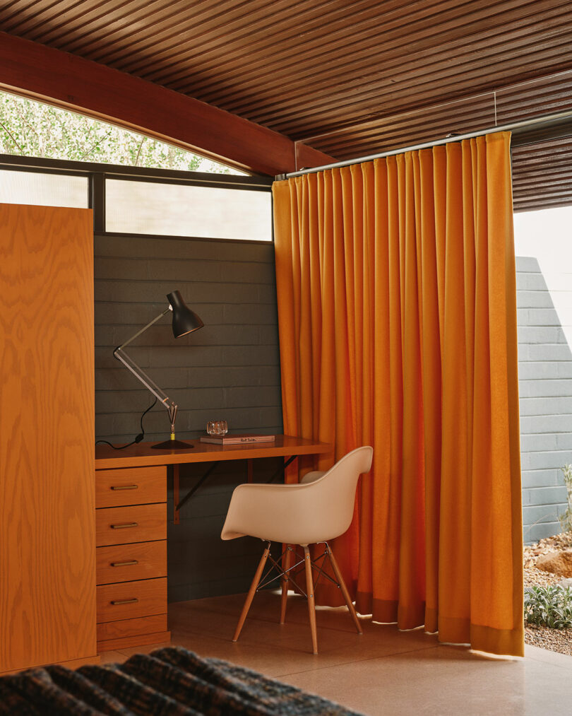 A wooden desk with drawers, a modern white chair, a black desk lamp, and orange curtains in a room with a wood ceiling and high windows.