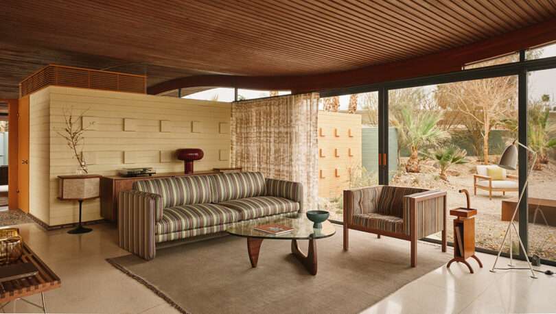 Mid-century modern living room with striped sofa, wooden armchair, glass coffee table, and floor-to-ceiling windows overlooking a desert garden.