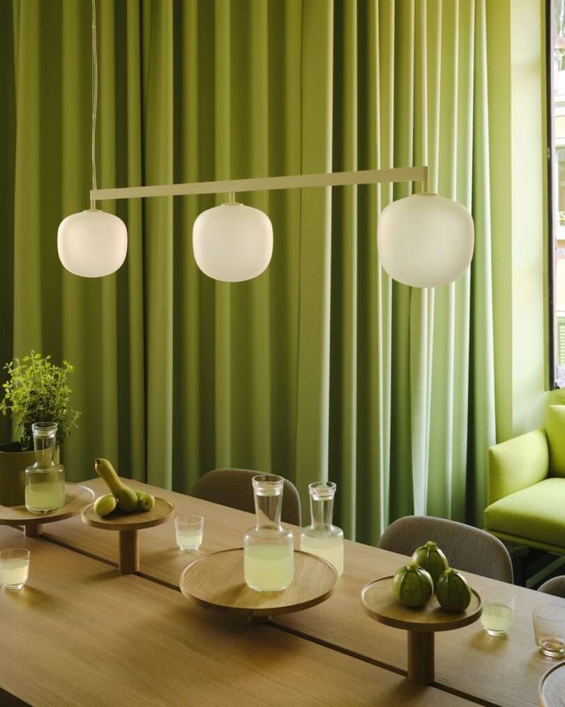 A modern dining table with wooden trays holding pears, figs, and a carafe of juice, under three globe pendant lights, with green curtains in the background