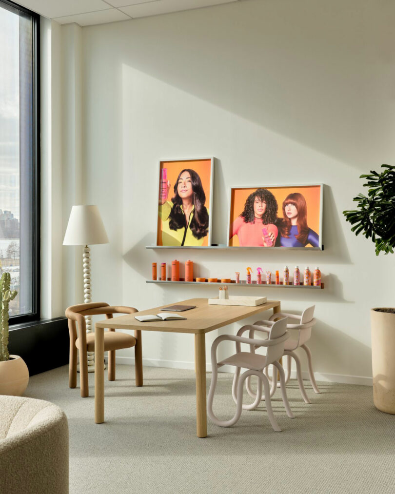 A modern office space with a wooden table, chairs, a potted plant, and two colorful posters of women with hair products on the wall above a shelf holding bottles.