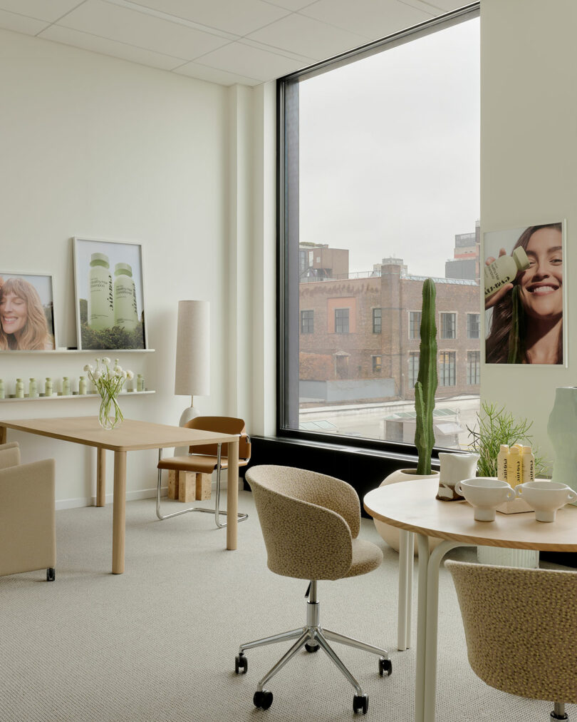 Modern office with neutral-toned furniture, a large window overlooking city buildings, potted plants, product displays, and posters featuring a smiling woman and skincare bottles.