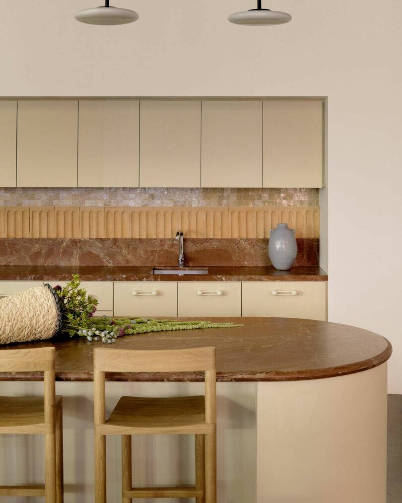 Modern kitchen with beige cabinets, a brown marble countertop, wooden stools, a vase, and greenery on the island; beige tiled backsplash and minimalist pendant lights above.