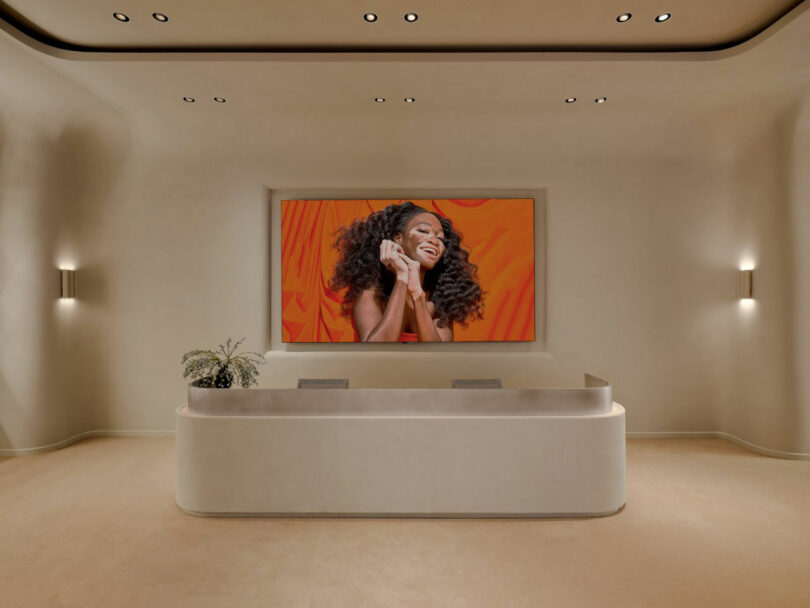Minimalist reception area with a curved desk, small plant, and a large wall photo of a smiling woman holding a phone, set against an orange backdrop.
