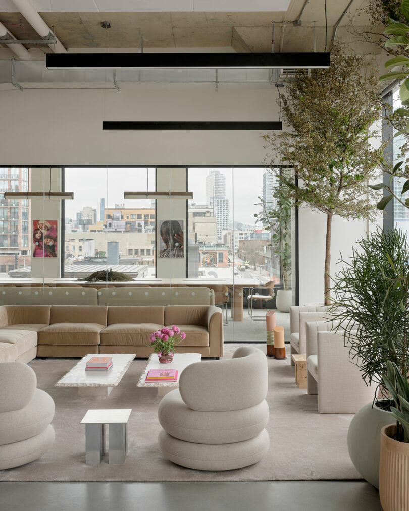 Modern lounge area with beige sofas and round chairs, large windows overlooking a cityscape, indoor plants, and contemporary decor elements.