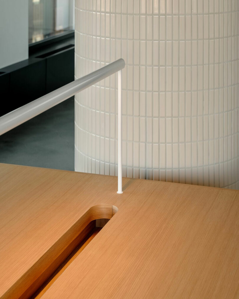 A white vertical railing passes through a wooden tabletop with a slot, set against a background of a large, tiled cylindrical column.