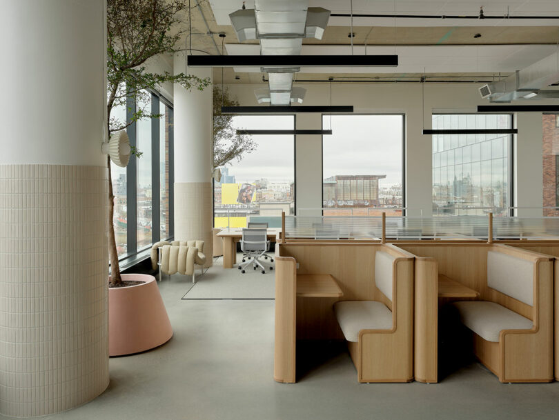 Modern office space with wooden booth seating, a desk with a chair near large windows, potted trees, and a city view outside. The interior has neutral colors and minimalist design.