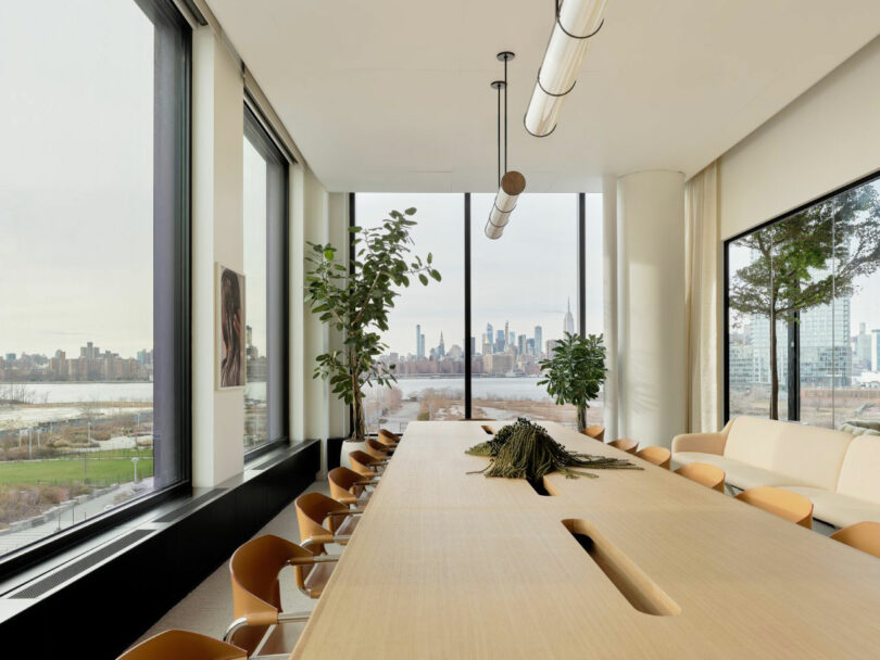 Modern conference room with a long wooden table, brown chairs, large windows overlooking a city skyline, indoor plants, and a neutral color palette.