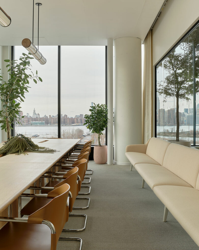 Modern conference room with a long table, brown chairs, beige sofa, large windows, indoor plants, and a city skyline view across a river.