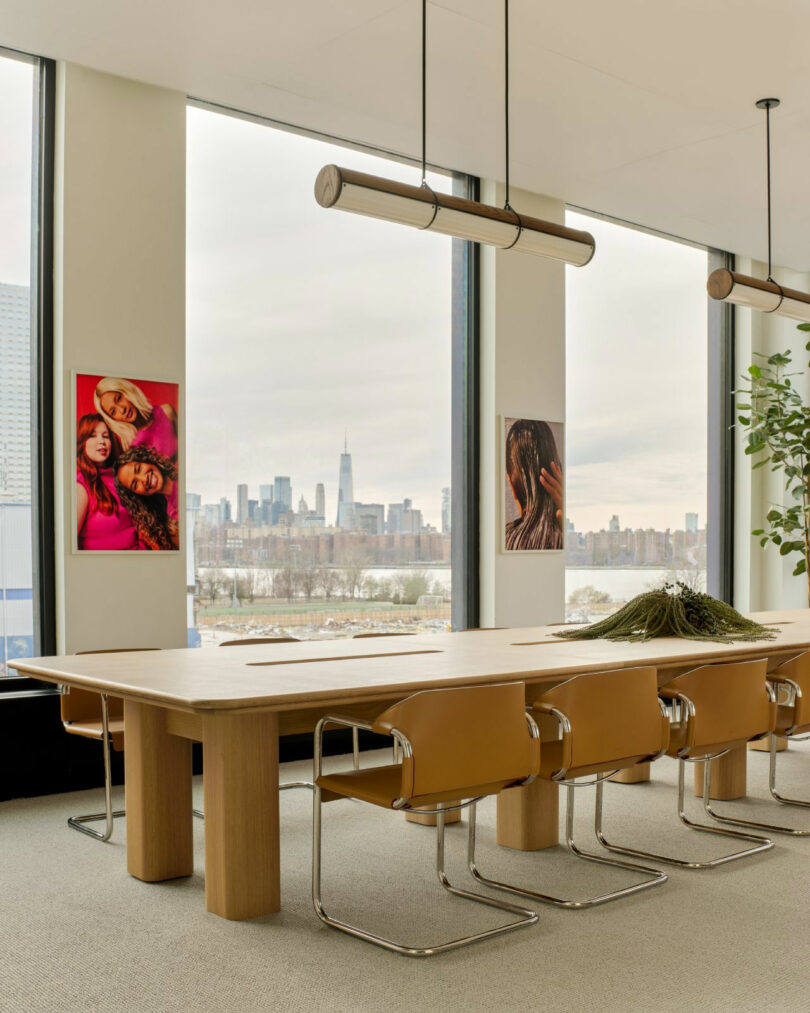 A modern conference room with tan chairs around a wooden table, large windows with a city skyline view, and colorful portraits on the walls.