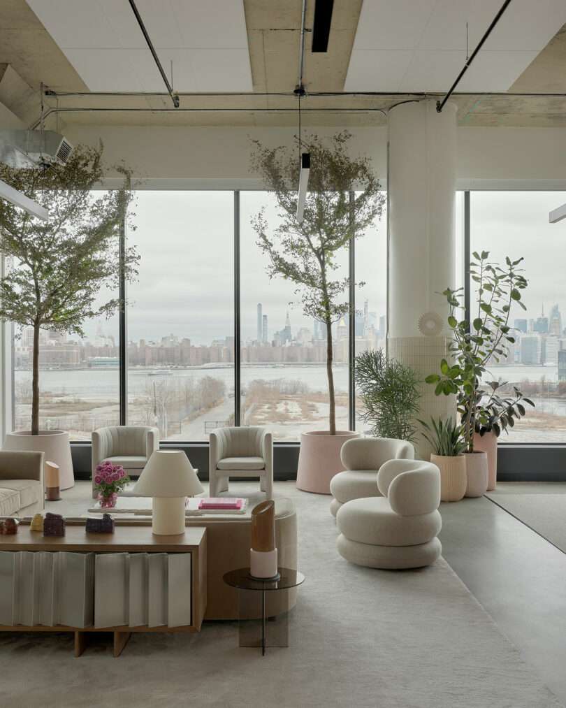 Modern office lounge with contemporary furniture, large windows, indoor plants, and a city skyline visible in the background.