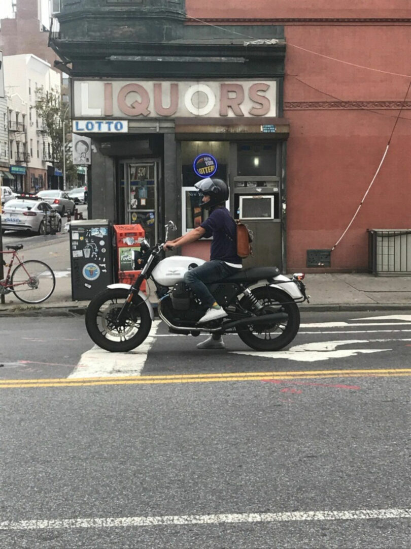 A person wearing a helmet and backpack sits on a white motorcycle stopped at a crosswalk in front of a liquor store, reminiscent of a scene designed by JUN AIZAKI.