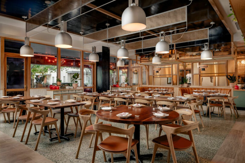 A modern, well-lit restaurant dining area designed by JUN AIZAKI, featuring wooden tables and chairs set with plates and napkins, and large windows along one side.