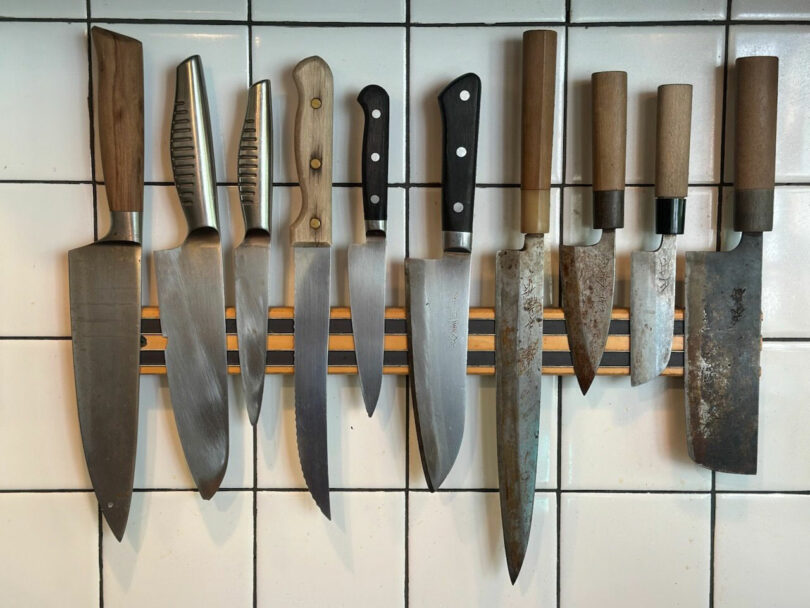 A row of various kitchen knives with different handle styles is mounted on a magnetic strip against a tiled wall, showcasing a sleek, JUN AIZAKI-inspired design aesthetic.