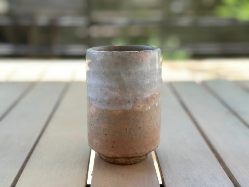 A handmade ceramic cup with a natural, earthy glaze—reminiscent of JUN AIZAKI's design philosophy—sits on a wooden table with a blurred background.