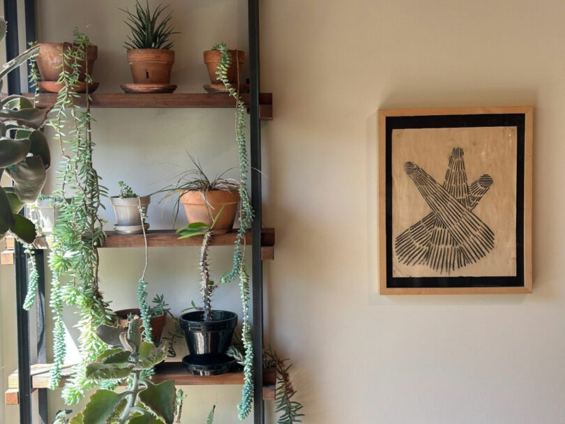 A wooden shelf with potted plants is next to a wall featuring a framed abstract black and white artwork in the style of JUN AIZAKI.