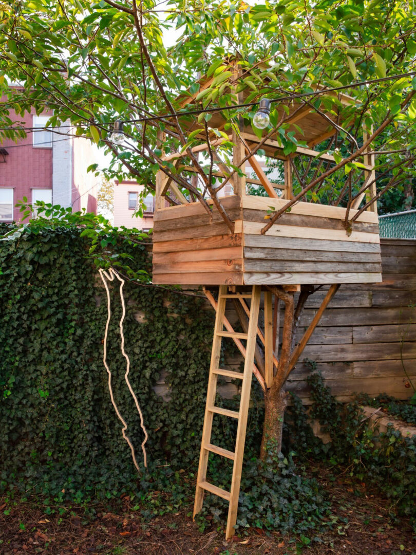 A wooden treehouse with a ladder, inspired by JUN AIZAKI’s natural designs, sits among the branches of a tree in a leafy backyard, next to a vine-covered wall and wooden fence.