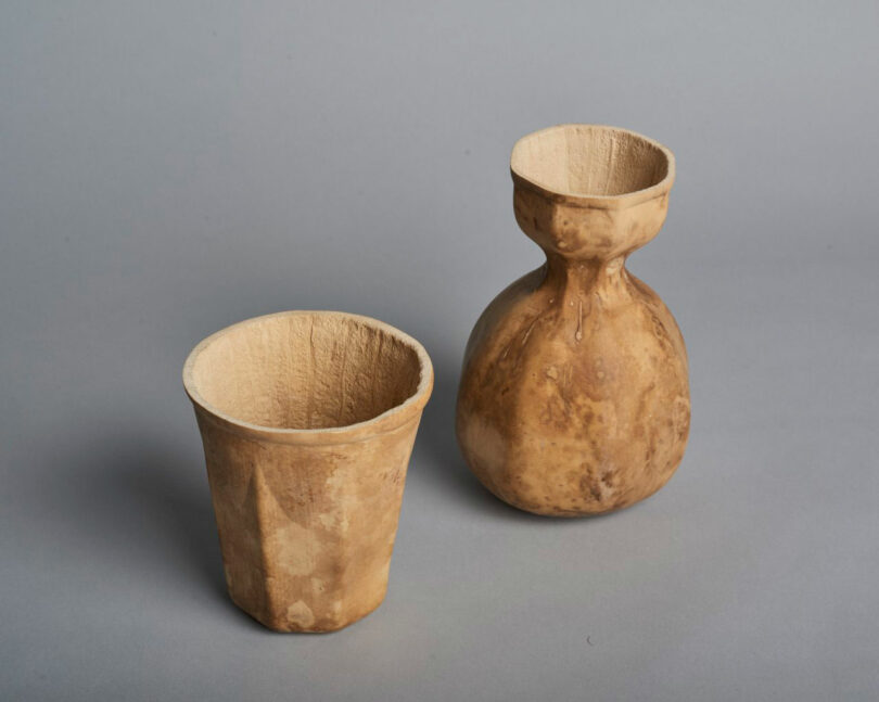 Two brown, rustic containers made from natural material—reminiscent of JUN AIZAKI's organic aesthetic—include a straight-sided cup and a round-bodied vessel with a narrow neck, both set on a plain gray background.