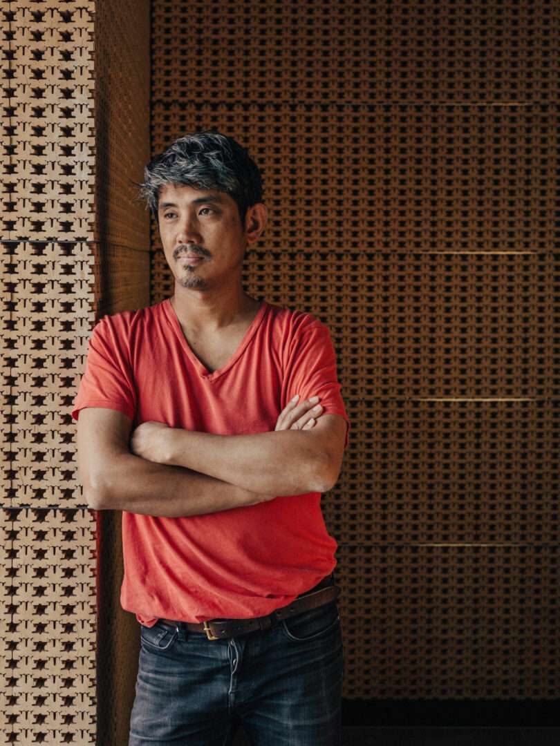 A man in a red T-shirt and jeans, resembling JUN AIZAKI, stands with arms crossed, leaning against a textured wall with a neutral expression.