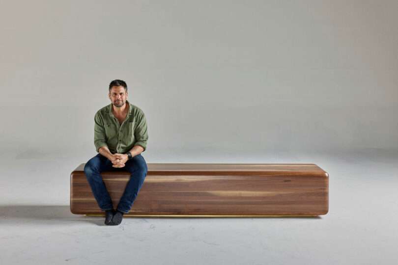 A man in a green shirt and jeans sits barefoot on a long, rectangular wooden bench in a plain, light-colored room.