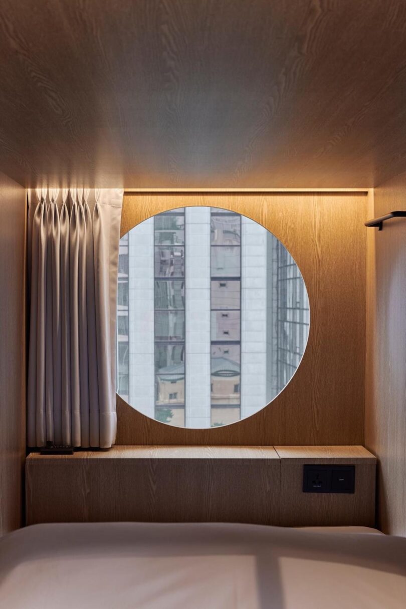 A round window on the other end of the capsule pod offers a city building view, set in a wooden wall above a bed; white curtains are drawn to one side and electrical outlets are visible below the window to the right