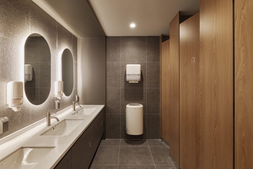 Modern public restroom for guests with capsule reservations at HOTEL DRIP&DROP featuring three wooden stall doors on the right, two sinks with mirrors above and wall-mounted soap dispensers to the left, a paper towel dispenser and trash can mounted on the back middle wall, and grey tiled floors and walls