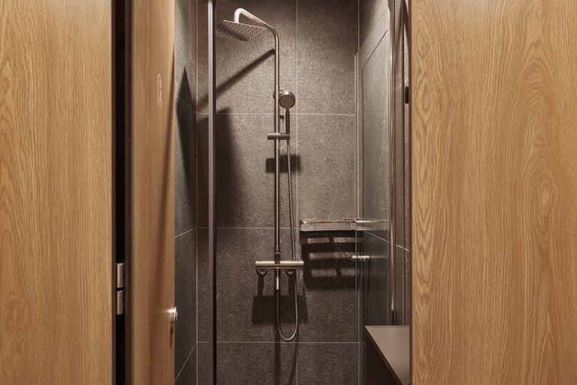 A modern hotel shower with a rainfall showerhead and glass door, surrounded by dark tiles and light wood doors