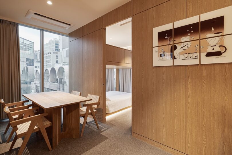 Modern hotel suite with wood-paneled walls, a dining table for four, large window, partial bedroom view, and framed coffee-themed art on the wall