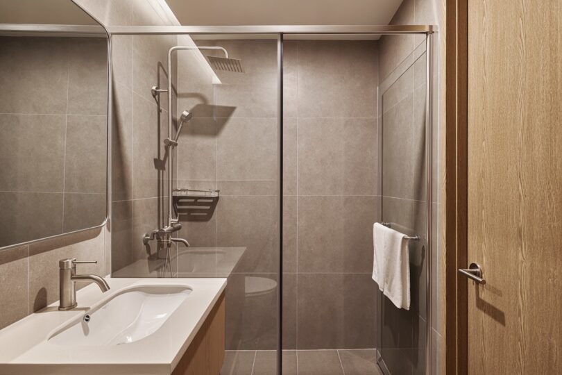 Modern hotel bathroom features a glass-enclosed shower, wall-mounted rainfall showerhead, grey tiles, rectangular sink, large mirror, and a towel hanging on a rail