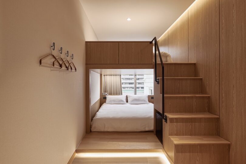 A compact modern bedroom at HOTEL DRIP&DROP features a double bed under a wooden loft, stairs on the right, and wall-mounted coat hooks on the left. Natural light streams through a window at the far end