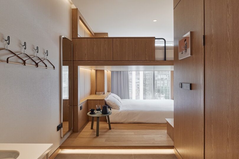 Modern HOTEL DRIP&DROP room with wooden furnishings, a loft bed above a larger bed, a small table with a kettle to make coffee, wall-mounted hooks, and a large window flooding the space with natural light