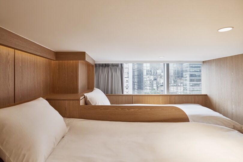 Two neatly made single beds with white linens are positioned in a small, wood-paneled loft at HOTEL DRIP&DROP, featuring a large window overlooking city buildings in the background.
