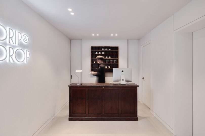 A person stands behind the dark wooden reception desk at HOTEL DRIP&DROP, with a computer, shelves of cups and coffee beans in containers behind, and