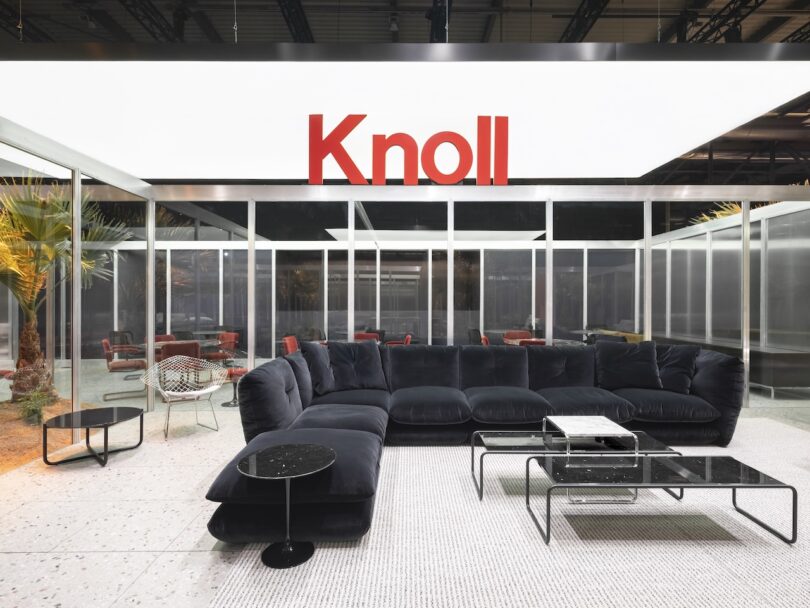 A modern showroom displays a large black sectional sofa with glass coffee tables under a bright sign reading 