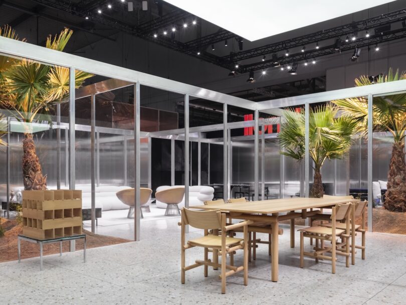 Modern indoor space with glass walls, palm trees, light wood dining table and chairs, white lounge chairs, and a terrazzo floor under bright ceiling lights