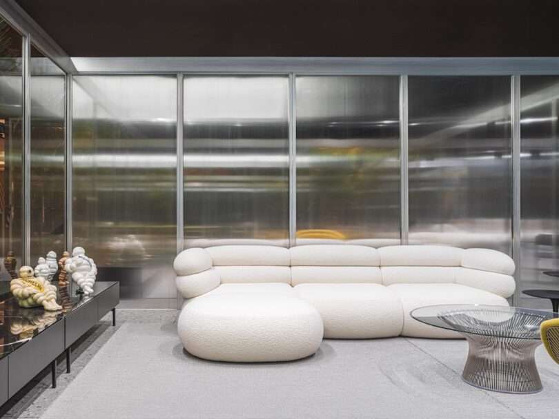 A modern living room with a white curved sectional sofa, glass coffee table, decorative sculptures on a sideboard, and frosted glass panel walls