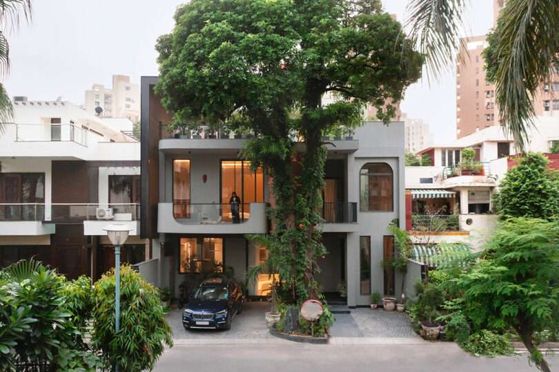Modern two-story house with large tree integrated into the front entrance, a car parked in the driveway, and a person standing on the balcony.