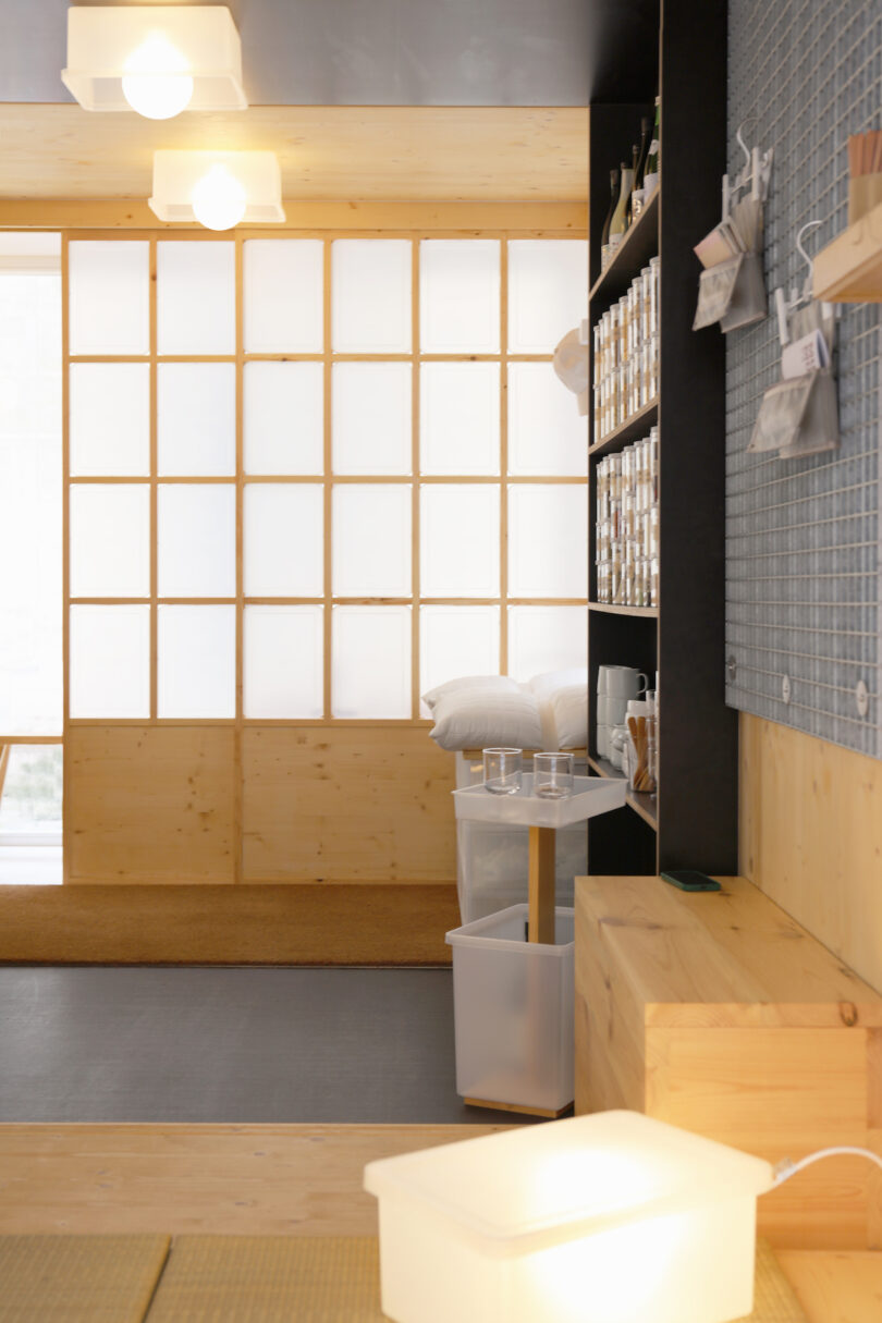 A modern, minimalist room with wooden furniture, frosted panel windows, shelves with jars, a trash bin, and soft overhead lighting