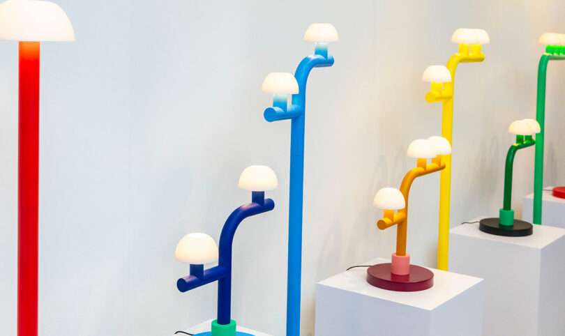 A display of colorful, modern lamps by Nader Gammas in red, blue, purple, yellow, and green, each with multiple white dome-shaped lights on white pedestals against a plain wall.